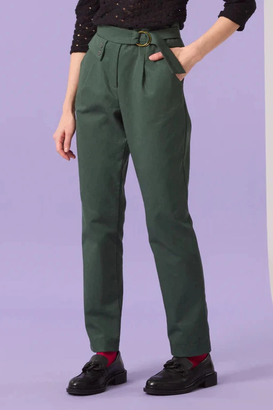 Waist-down view of a woman wearing the Saules Pants by Cokluch in Eden, with a fly closure, an attached D-ring belt, a button and tab detail, side pockets, and an ankle length, made from OEKO-TEX certified fabric. She is wearing them with a black top and standing in front of a purple background. 