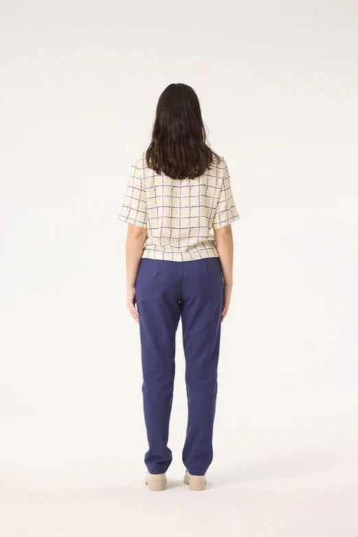 Back view of a woman wearing the Saules Pants by Cokluch in Twilight with a fly closure, an attached D-ring belt, a button and tab detail, side pockets, and an ankle length, made from OEKO-TEX certified fabric. She is wearing them with the Sideral Top in Cream Plaid and standing in front of a white background. 