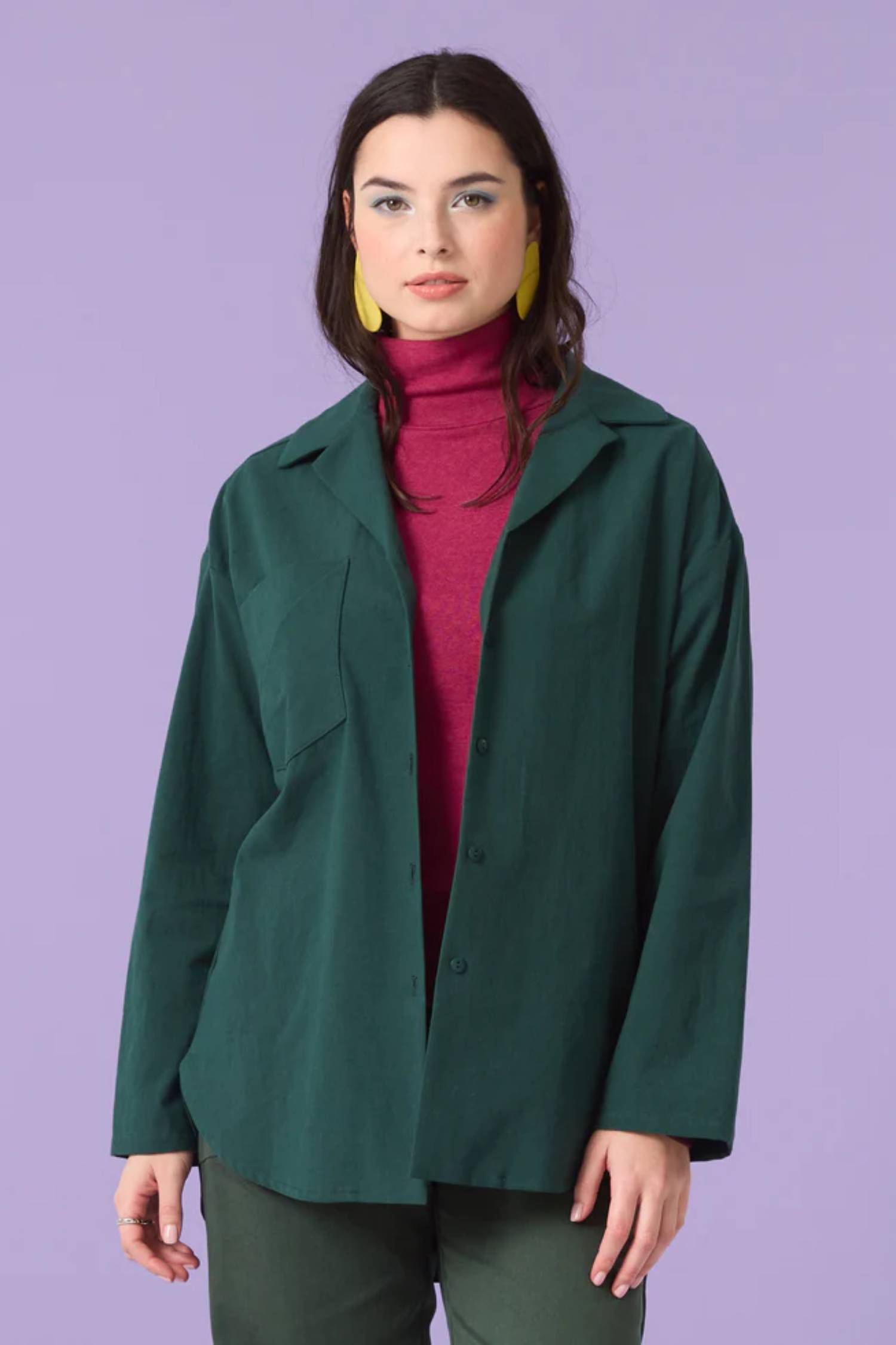 3/4 shot of a woman wearing the Willow Top by Cokluch in Eden over a pink turtleneck, standing in front of a purple background 