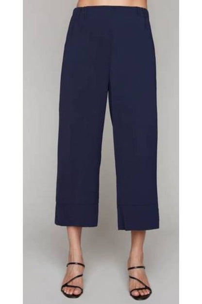 A waist-down view of a woman wearing the Opal Pant by Compli K in Navy, with a high waist, wide cropped legs, and a button detail on the cuffs. She's standing in front of a grey background. 