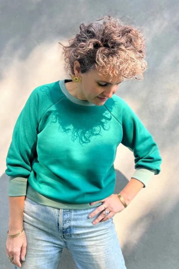 A woman wearing the Lipa Crewneck Sweatshirt by Kazak in Cedar and Sage, featuring a round neck, 3/4 raglan sleeves, and a fitted band across the bottom. She is wearing it with jeans and is standing outside in front of a grey wall.