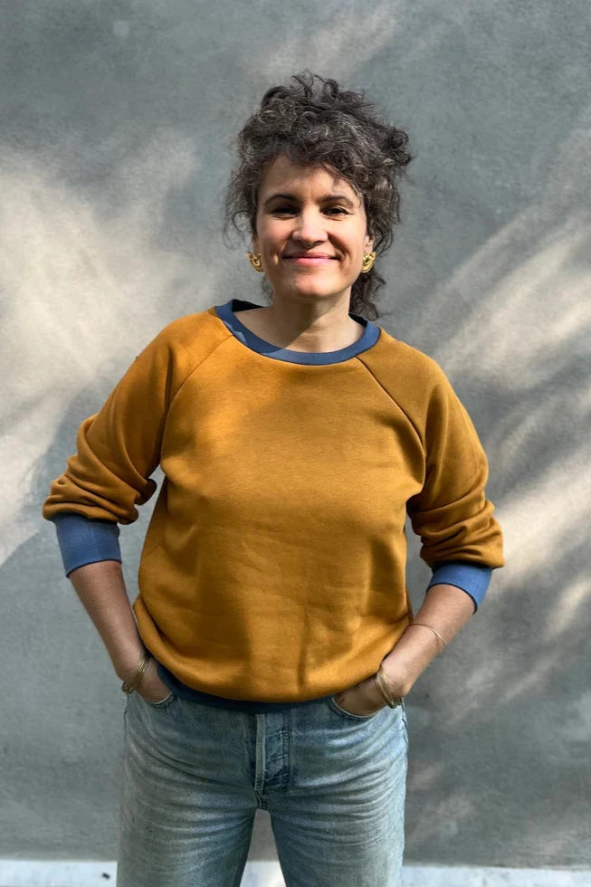 A woman wearing the Lipa Crewneck Sweatshirt by Kazak in Mustard and Steel, featuring a round neck, 3/4 raglan sleeves, and a fitted band across the bottom. She is wearing it with jeans and is standing outside in front of a grey wall.