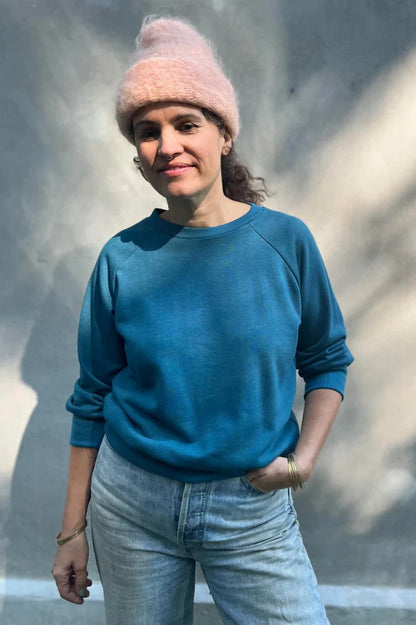 A woman wearing the Lipa Crewneck Sweatshirt by Kazak in Tealfeaturing a round neck, 3/4 raglan sleeves, and a fitted band across the bottom. She is wearing it with jeans and a pink tuque and is standing outside in front of a grey wall  