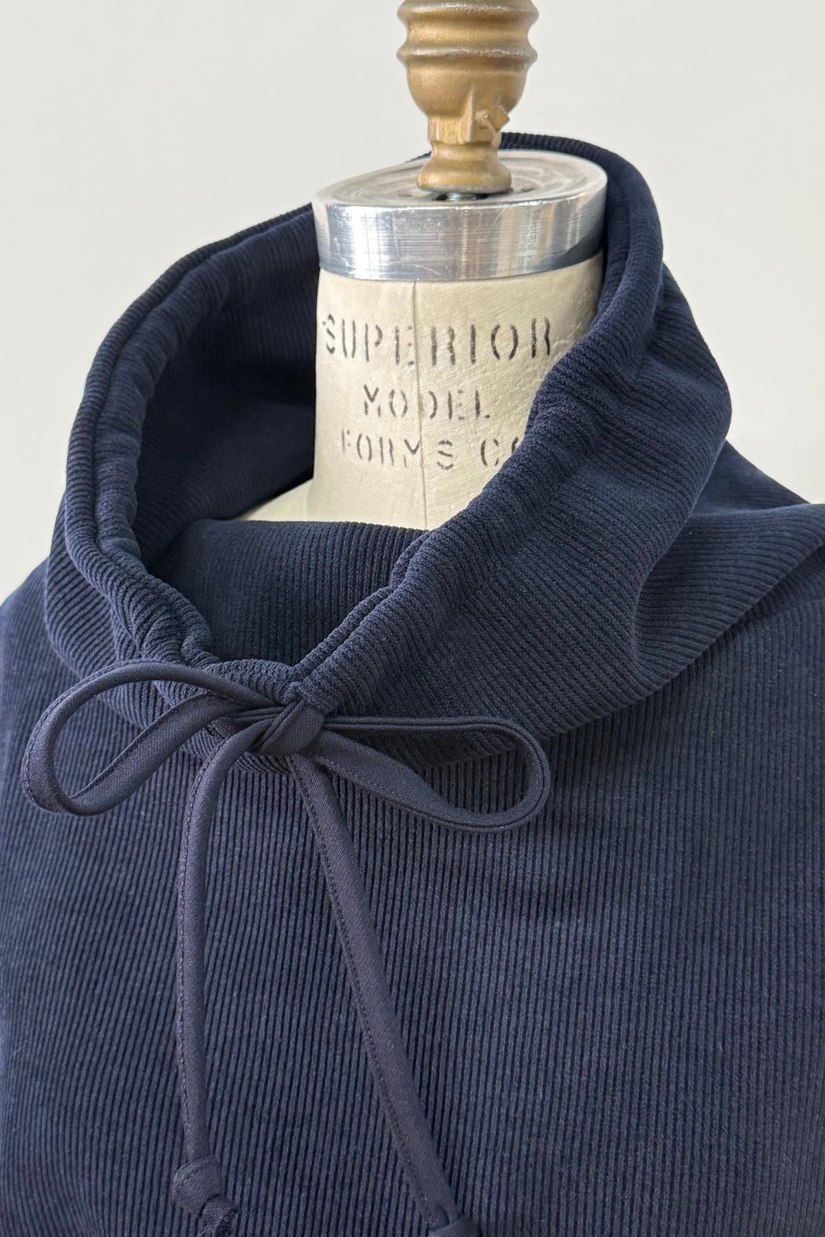 A close up view of the Ramos Sweater by Kollontai in a ribbed navy corduroy-like fabric, with a large cowl neck with a drawstring and a kangaroo pocket, is shown on a mannequin against a white background. 