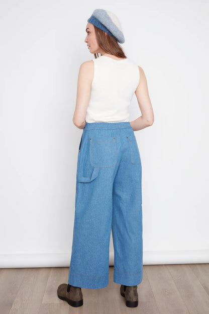 Back view of a woman wearing the Brooke Wide Leg Denim Pants by Julei in light denimm, with large patch pockets, a zipper, elastic at the back waist, and a carpenter loop. She is wearing them with a white top and standing in front of a white background. 