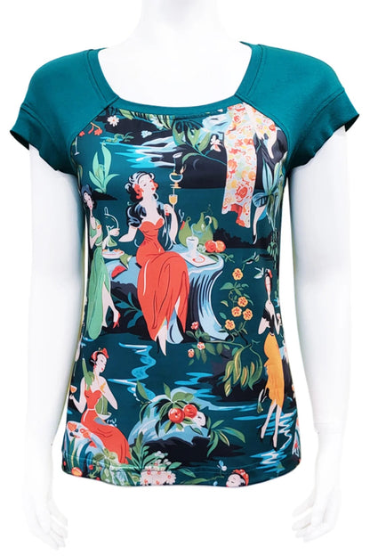 The Decades Era Tee by Mandala in 40s Glam, featuring a print with a tropical scene and pinup girls and contrasting teal shoulders and back, is shown on a mannequin in front of a white background. 