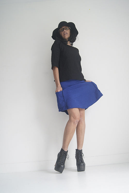 A woman wearing the Oversized Top by SI Design in Black, featuring a scoop neck, elbow-length sleeves, and a slightly relaxed fit. She is wearing with a blue skirt and standing in front of a grey background. 