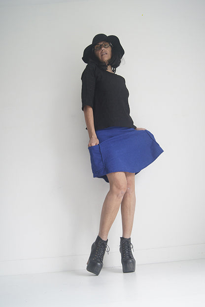 A woman wearing the Oversized Top by SI Design in Black, featuring a scoop neck, elbow-length sleeves, and a slightly relaxed fit. She is wearing with a blue skirt and standing in front of a grey background. 