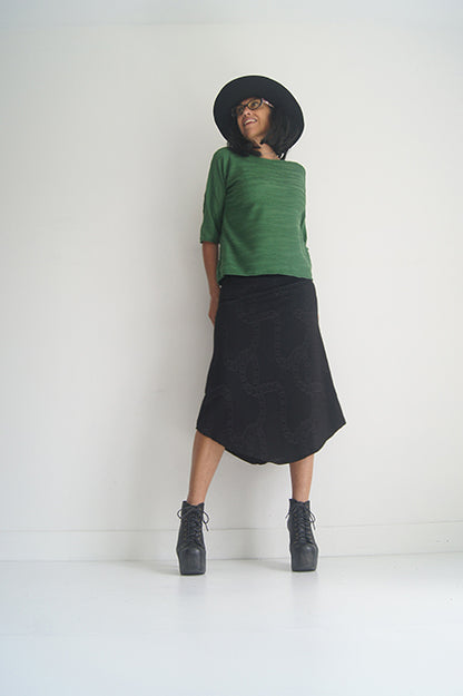 A woman wearing the Oversized Top by SI Design in Green, featuring a scoop neck, elbow-length sleeves, and a slightly relaxed fit. She is wearing with a black skirt and standing in front of a grey background. 