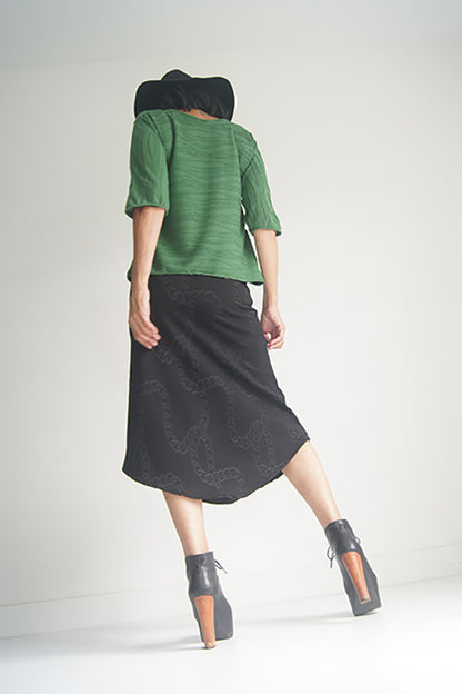 Back view of a woman wearing the Oversized Top by SI Design in Green, featuring a scoop neck, elbow-length sleeves, and a slightly relaxed fit. She is wearing with a black skirt and standing in front of a grey background. 