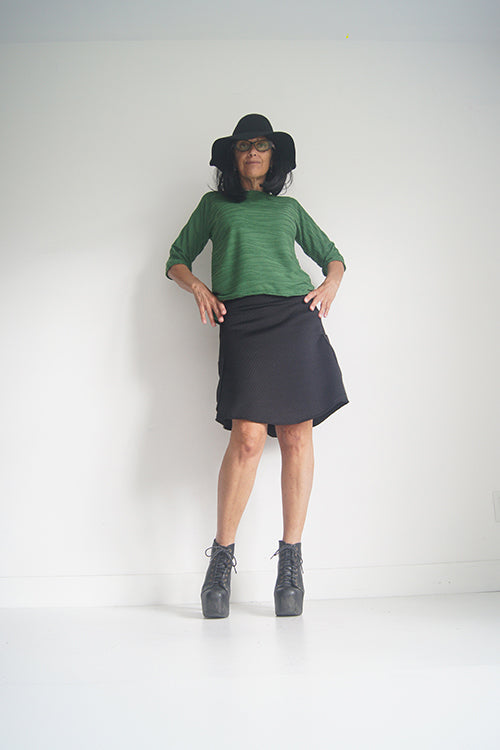 A woman wearing the Oversized Top by SI Design in Green, featuring a scoop neck, elbow-length sleeves, and a slightly relaxed fit. She is wearing with a black skirt and standing in front of a grey background. 