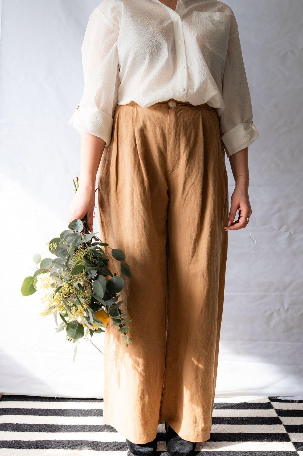 Neck-down view of a woman wearing the Ania Blouse by Cokluch in Off-White and beige pants holding a bouquet of flowers on a striped floor.