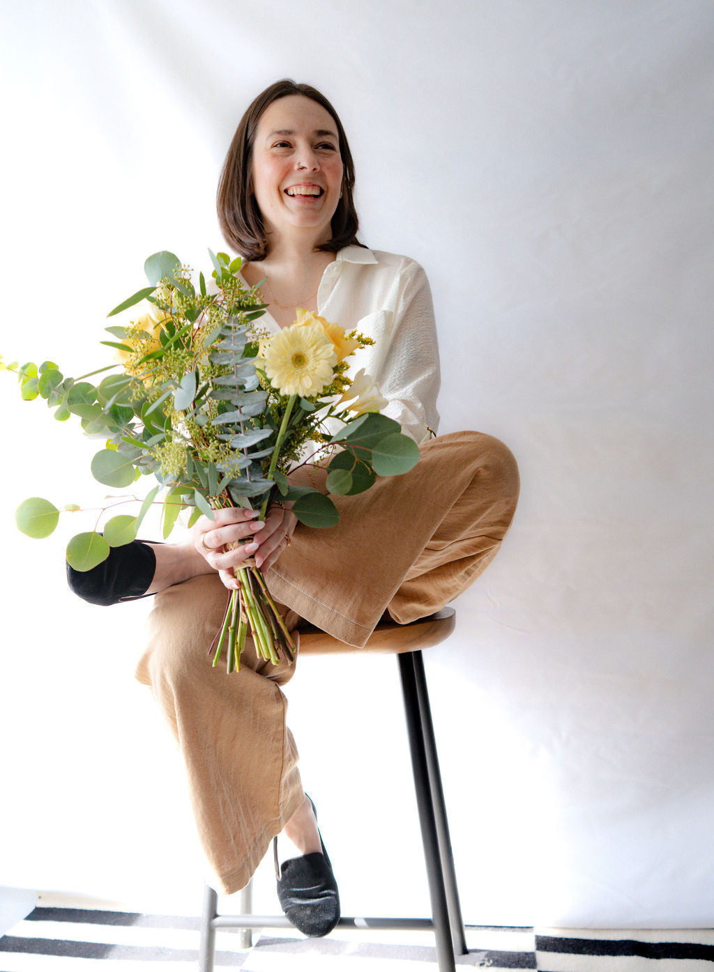 Woman wearing the Ania Blouse by Cokluch in Off-White, holding a bouquet of flowers sitting on a stool against a white wall