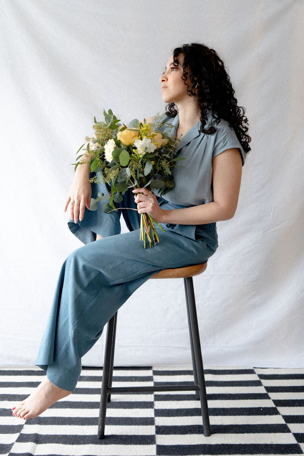 A woman wearing the Hemera Shirt by Cokluch in Sky, with a V-neck collar, button front, short extended sleeves, and a relaxed fit. She is wearing it with blue pants and sitting on a stool in front of a white background. 