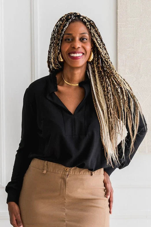A woman wearing the Sweatshirt Blouse by Dotty in Black, featuring a ruffled v-neck, long sleeves with banded cuffs, and a relaxed fit. She is wearing it with a tan skirt and standing in front of a white wall. 