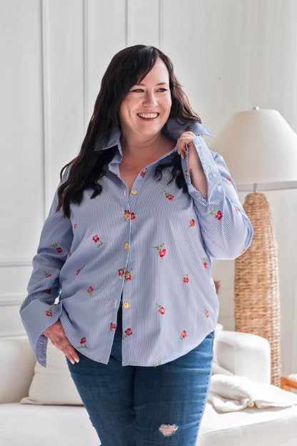 A woman wearing the Stripes and Roses shirt by Dotty in navy/white stripe with embroidered red roses, a collar, vintage gold buttons up the front, and double button cuffs. She is wearing it with jeans and standing in front of a white sofa. 