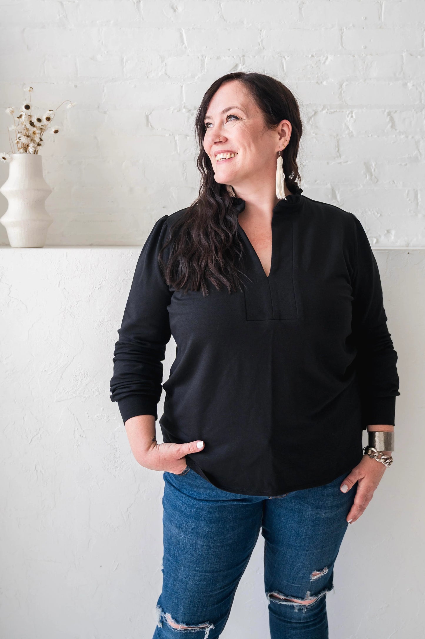 A woman wearing the Sweatshirt Blouse by Dotty in Black, featuring a ruffled v-neck, long sleeves with banded cuffs, and a relaxed fit. She is wearing it with jeans and standing in front of a white wall. 