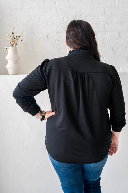 Back view of a woman wearing the Sweatshirt Blouse by Dotty in Black, featuring a ruffled v-neck, long sleeves with banded cuffs, and a relaxed fit. She is wearing it with jeans and standing in front of a white wall. 