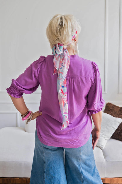 Back view of a woman wearing the Orchid Blouse by Dotty in Magenta, with a V-neck with a banded collar and elbow length smocked sleeves. She is wearing it with jeans standing in front of a couch. 