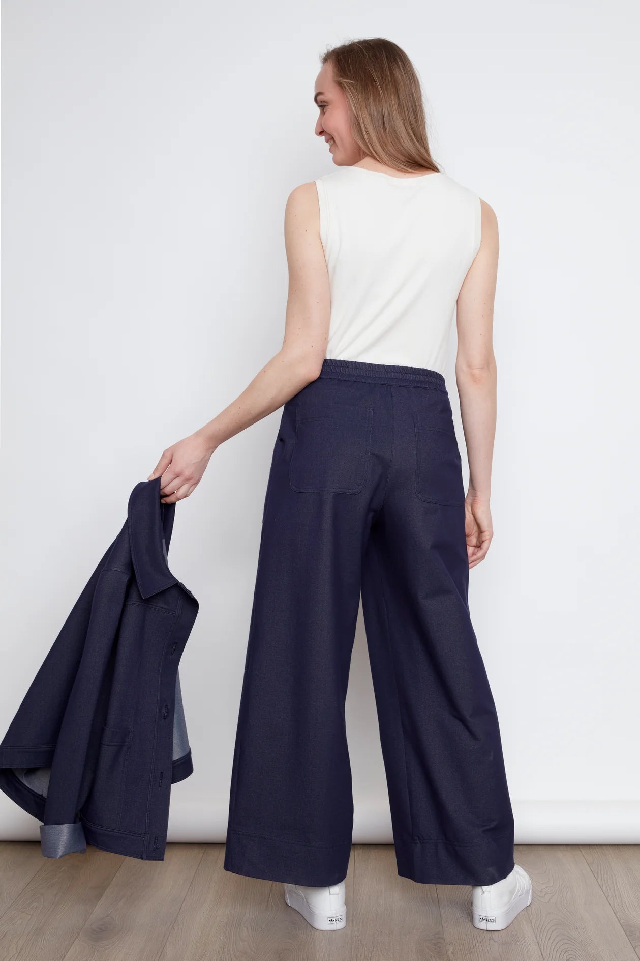 Back view of a woman wearing the Lucille Tank by Julei in Natural White, a ribbed tank with a round neck. She is wearing it with denim pants and holding a denim jacket in her hand. 