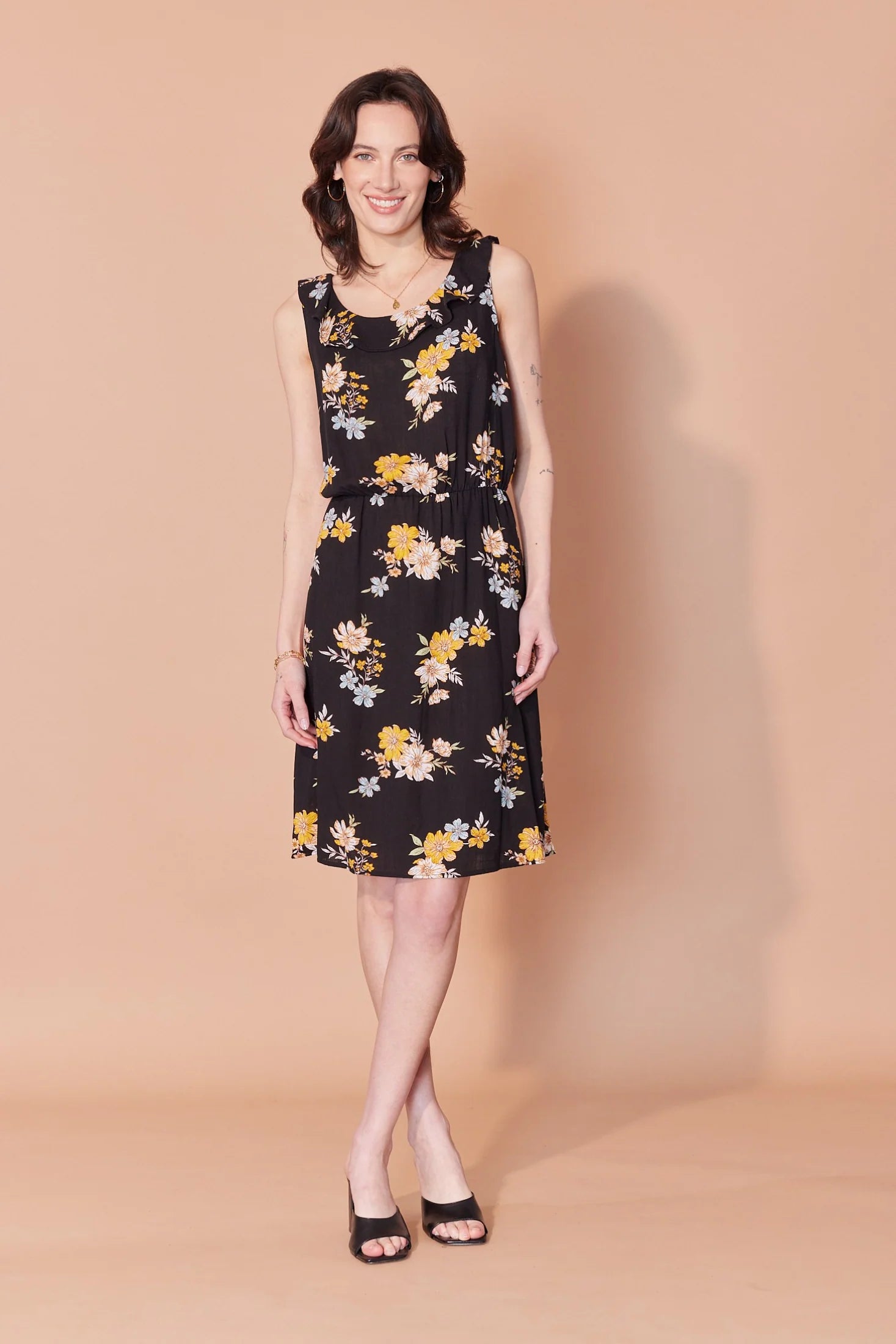 A woman wearing the Juliette Dress by Cherry Bobin in Floral print on black, a sleeveless dress with a ruffle at the neck, elastic wait, and knee length skirt. She is standing against a beige background