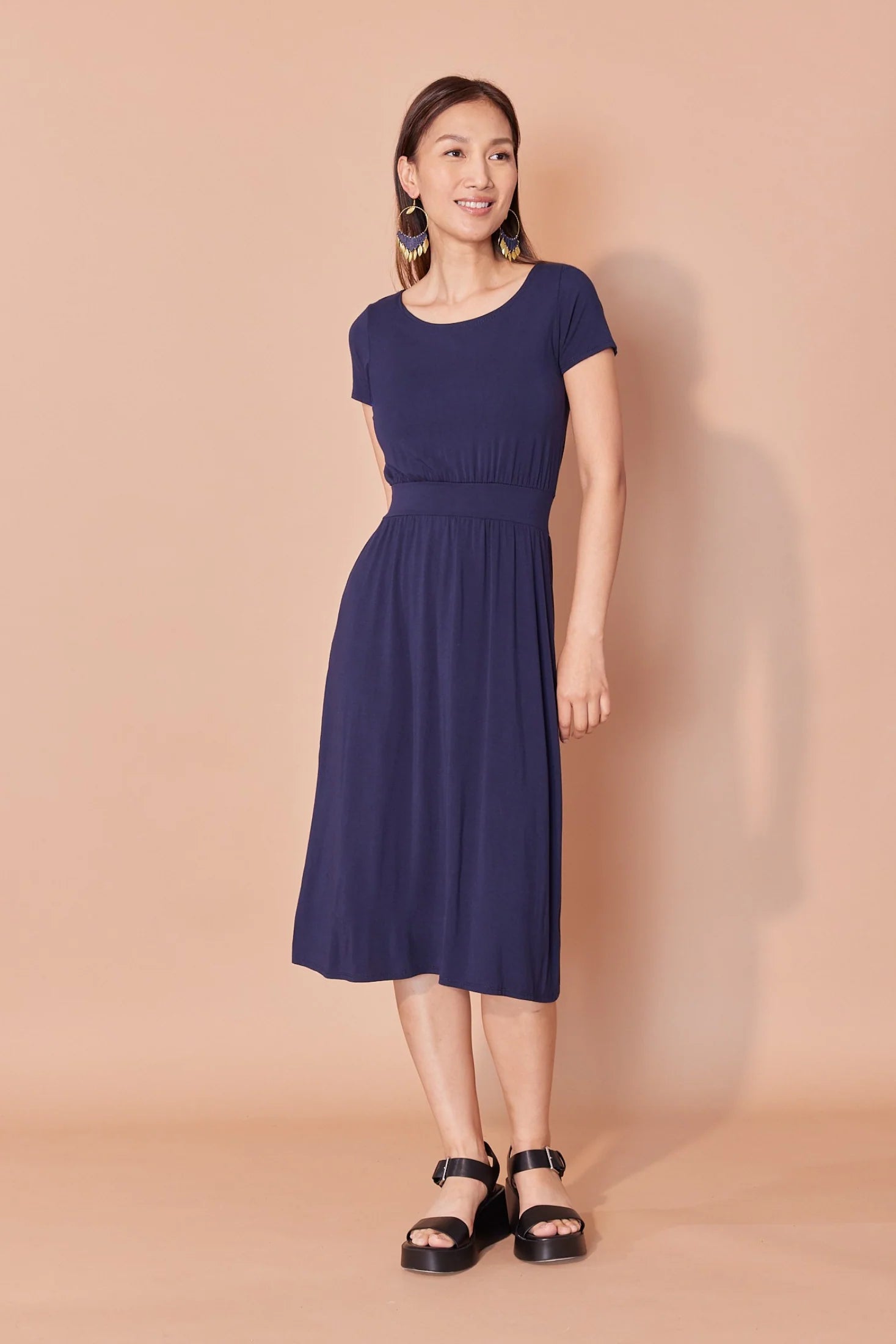 A woman wearing the Amelie Dress by Cherry Bobin in Navy, a below-the-knee dress with a fit and flare shape and short sleeves. She is standing against a beige background