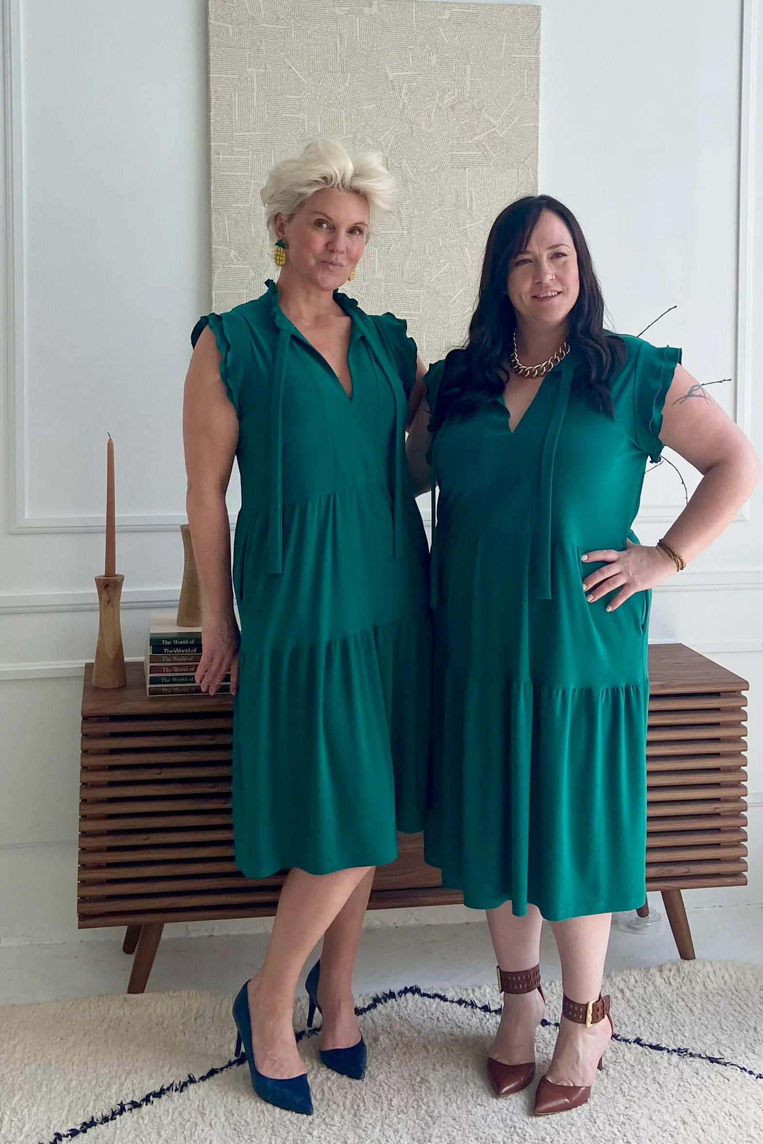Two women wearing the Ruffle Sleeve Tie Dress by Dotty, in emerald green, standing in front of a low table