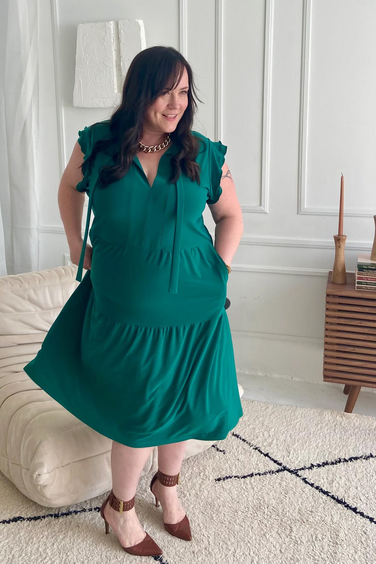 A woman twirling in the Ruffle Sleeve Tie Dress by Dotty, in emerald green, standing in front of a chair