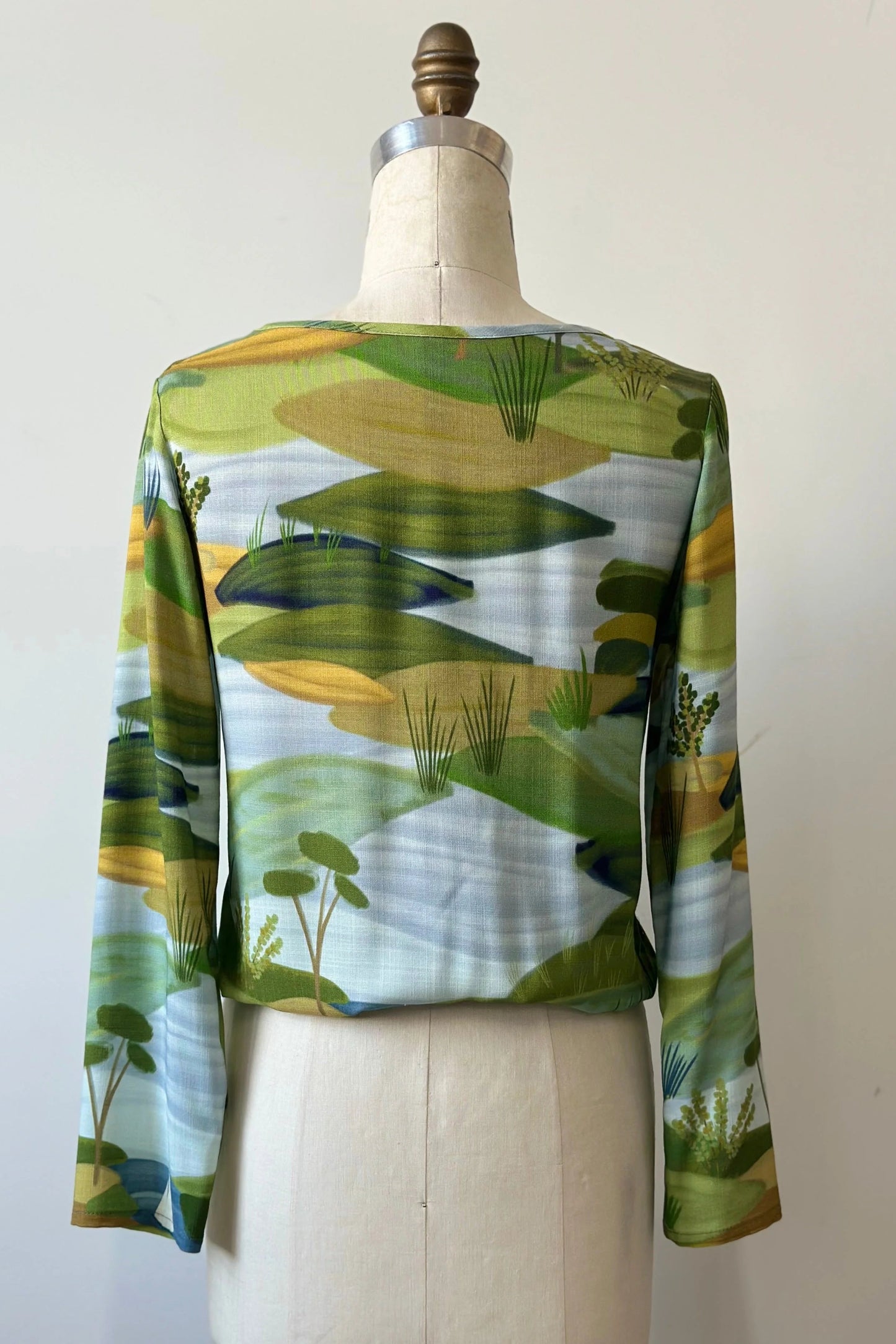A back view of the Belica Blouse by Kollontai in a Granny Smith toned nature print, with a wide neck, long sleeves, linen-look fabric, and an elasticized waist for a subtle blouson effect, is shown on a mannequin in front of a white background. 