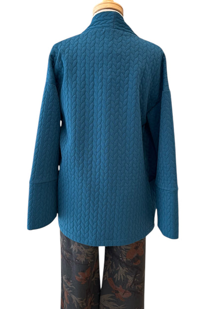 A back view of the Ina Coatigan by Tangente in Peacock, an open front cardigan in a cable-knit textured fabric, with dropped sleeves and patch pockets, is shown on a mannequin against a white background. 