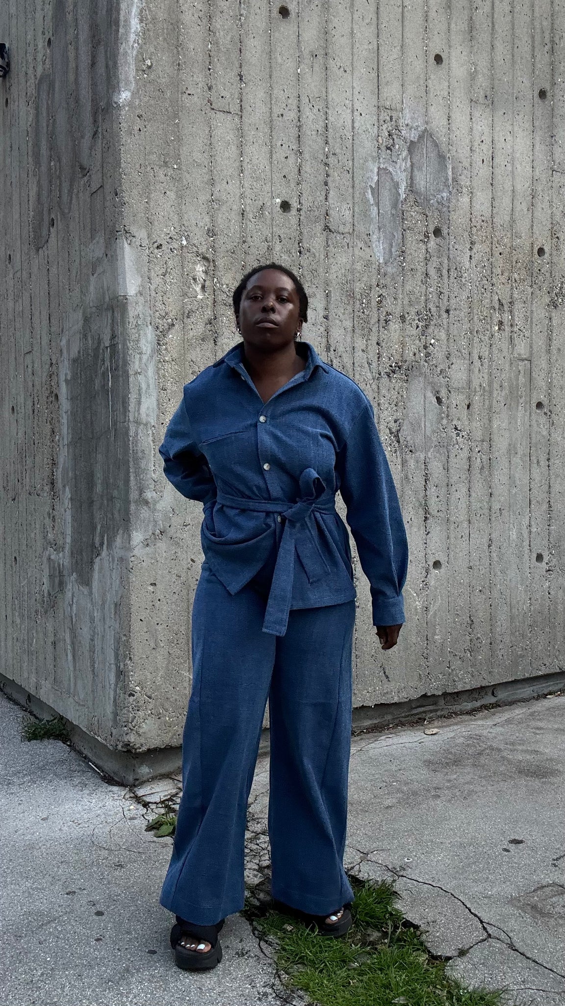  A woman wearing the Vienna Pants by MAS in Denim,  wide legged pants with a high waist and a zip front. She is wearing them with a matching jacket and standing outside by a cement building. 