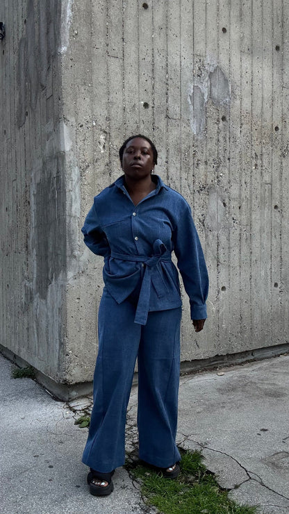  A woman wearing the Vienna Pants by MAS in Denim,  wide legged pants with a high waist and a zip front. She is wearing them with a matching jacket and standing outside by a cement building. 