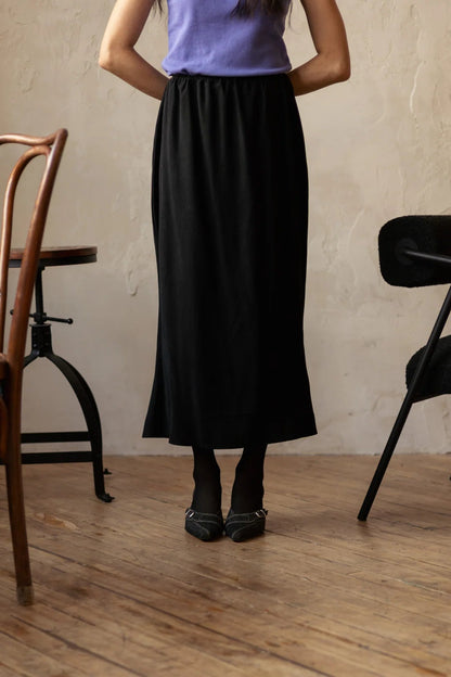 Chest-down view of a woman wearing the Simone Skirt by Lights of All in Black, a sleek calf-length skirt with a flared hem and an elastic waist. She is wearing it with a purple top and standing in a room with furniture. 