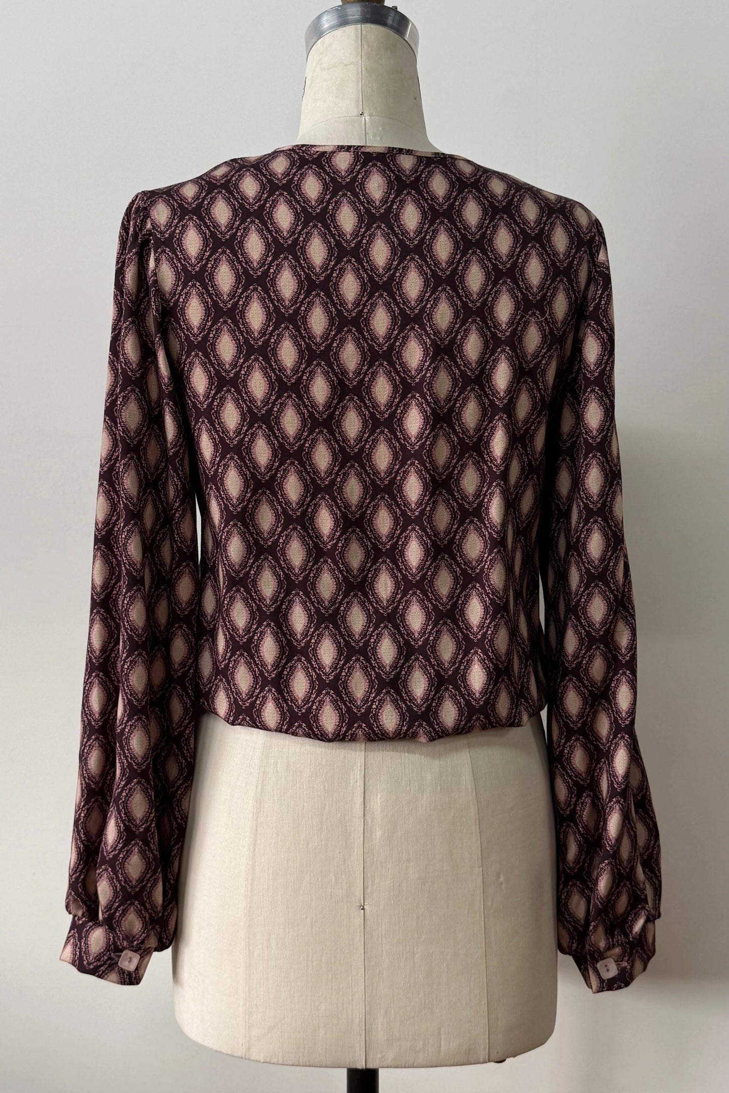 A back view of the Isabella Blouse by Kollontai, a burgundy and pink patterned blouse with a tie detail at the neck, balloon sleeves with square jewel buttons, and an elastic waist, is shown on a mannequin against a white background. 