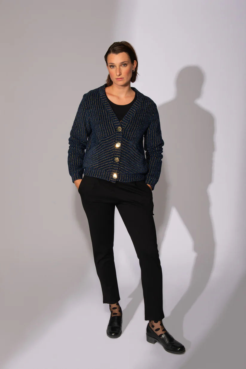 A woman wearing the Gold Lion Knit Cardigan by Eve Lavoie in Navy, featuring a V-neck, gold thread embedded in the navy fabric, gold buttons, and bias pockets. She is wearing it with black pants and standing in front of a white background. 