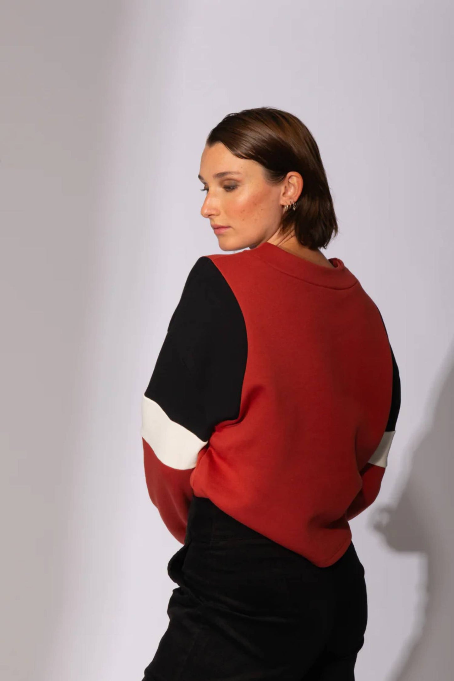 Back view of a woman wearing the Eve Lavoie Sour Time crewneck in Terracotta with Tricolour Cutout, featuring black/terracotta/white colourblocking. She is wearing black pants and standing in front of a grey background 