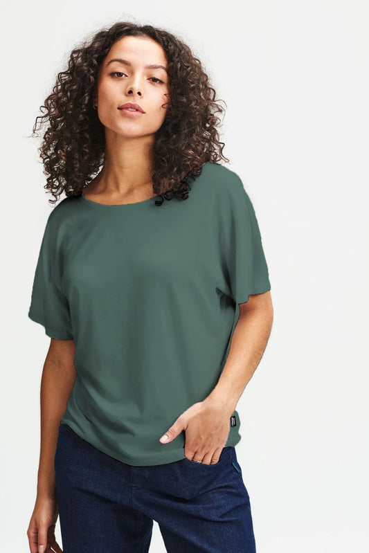 A woman wearing the Betty Shirt by  Message Factory in Green, a cotton/Tencel tee with a wide neck and short extended sleeves. She is wearing it with jeans and standing in front of a white background. 