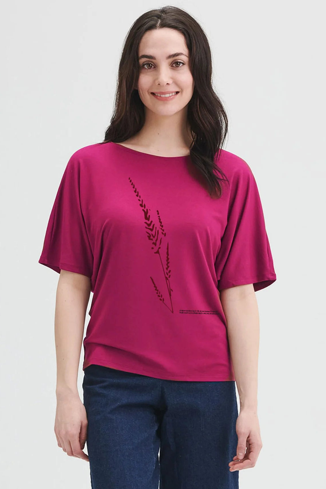A woman wearing the Betty Top by Message Factory in Raspberry, a bamboo top with short extended sleeves, a plant print, and French inscription about nature. She is wearing it with jeans and standing in front of a white background.