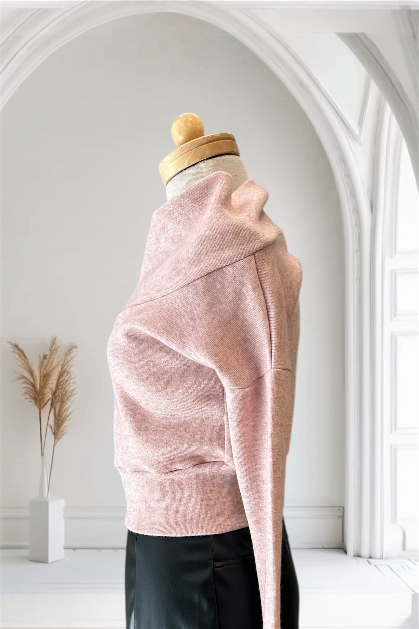 A side view of the Changes Sweater by Allison Wonderland in Pink, featuring a large cowl neck, long dropped sleeves, and a wide waistband, is shown with the Desire Skirt in Black against a white arched backdrop. 