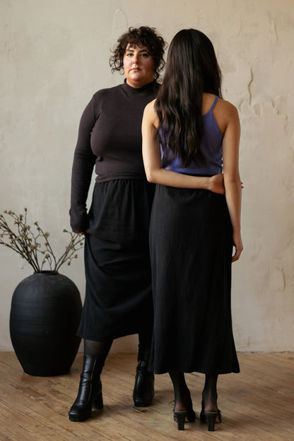 Two woman wearing the Simone Skirt by Lights of All in Black, a sleek calf-length skirt with a flared hem and an elastic waist. One is facing backwards and is wearing it with a purple top, the other is facing forwards and is wearing it with a brown turtleneck. They are standing in a room with furniture. 