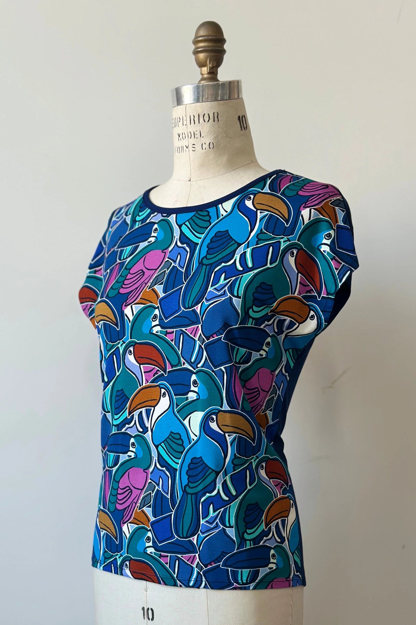 The Hanni Top by Kollontai in Admiral, a cap sleeved relaxed top with a jewel-toned parrot pattern on the front a solid blue back, is shown on a mannequin in front of a white background. 