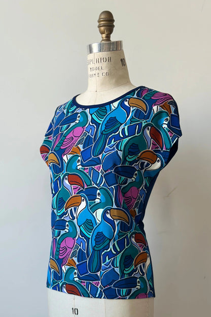 The Hanni Top by Kollontai in Admiral, a cap sleeved relaxed top with a jewel-toned parrot pattern on the front a solid blue back, is shown on a mannequin in front of a white background. 