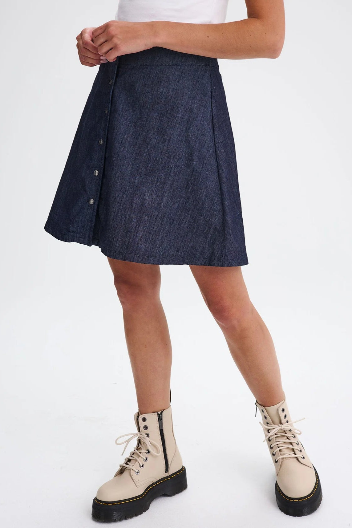 Waist down view of a woman wearing the Marilyn Denim Skirt by Message Factory, an A-line mini-skirt with a button front. She is wearing it with a white top and is standing in front of a white background. 