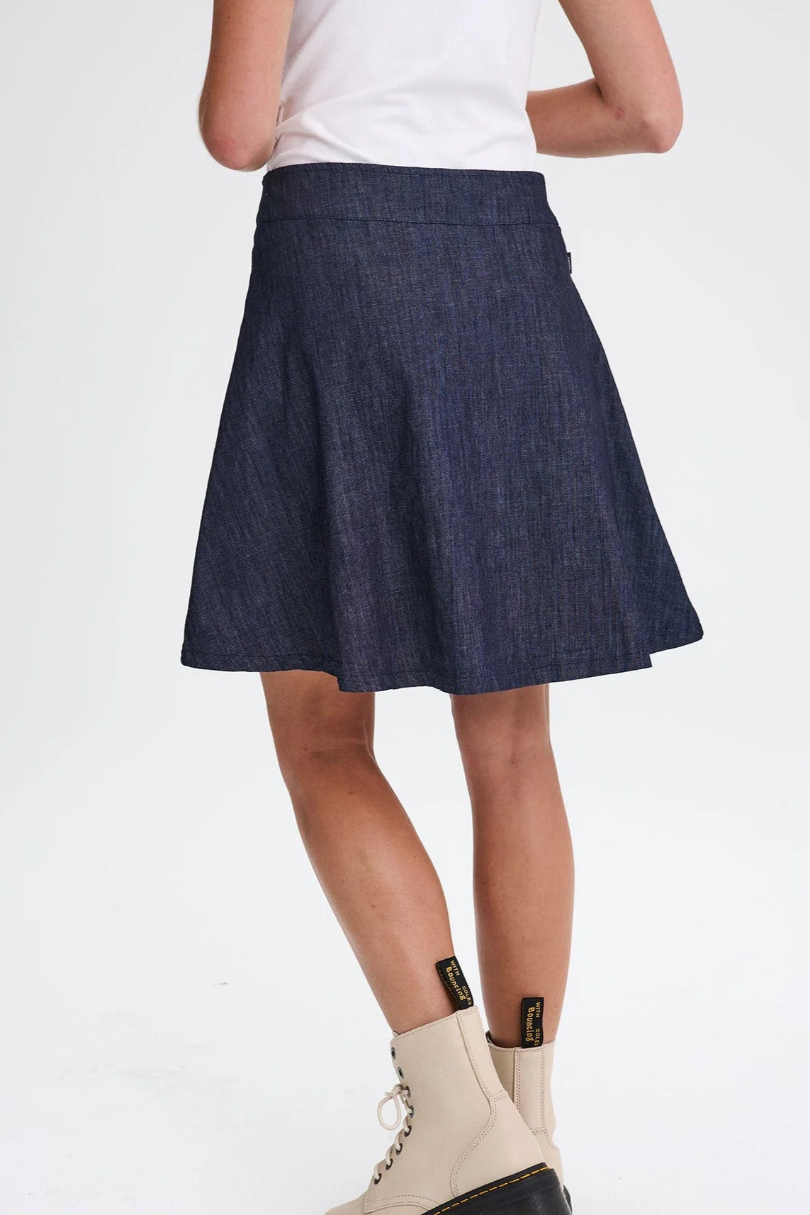 Back waist down view of a woman wearing the Marilyn Denim Skirt by Message Factory, an A-line mini-skirt with a button front. She is wearing it with a white top and is standing in front of a white background. 