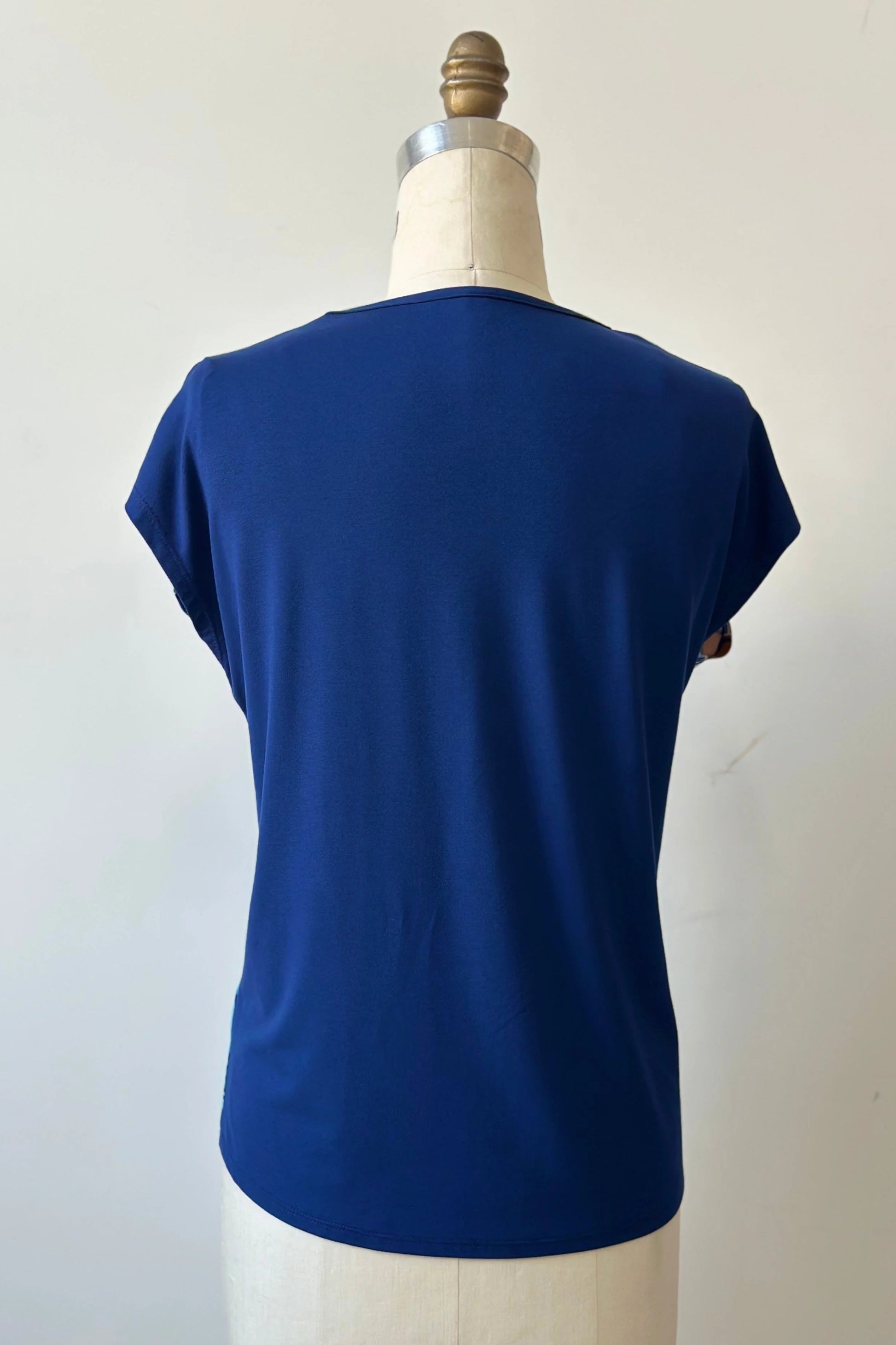 Back view of the Hanni Top by Kollontai in Admiral, a cap sleeved relaxed top with a jewel-toned parrot pattern on the front a solid blue back, is shown on a mannequin in front of a white background. 