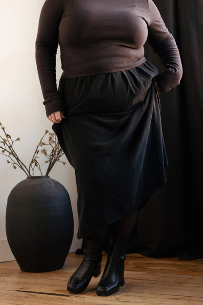 Neck down view of a woman wearing the Simone Skirt by Lights of All in Black, a sleek calf-length skirt with a flared hem and an elastic waist. She is wearing it with a brown turtleneck and standing in a white room by a black curtain. 
