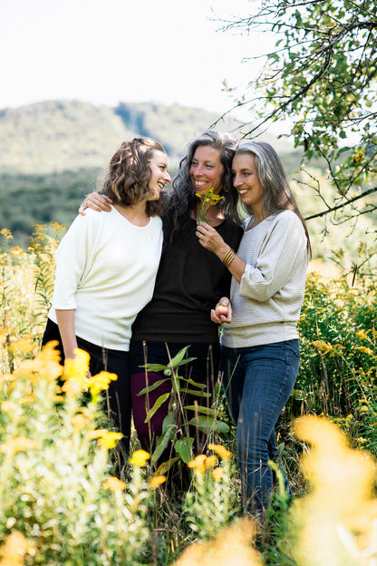 Three women wearing the Perla Top by Marie C,  one in Ivory and one in Black, with a wide round neck, 3/4 batwing sleeves, and fitted waist in ribbed knit fabric. They are standing outside. 