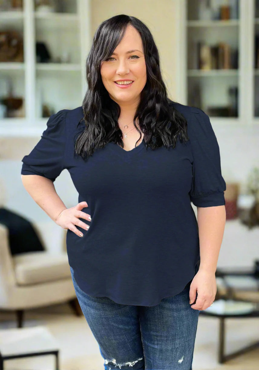 A woman wearing the FW Darling Tee by Dotty in Navy, featuring a v-neck, pleated shoulders, and elbow length sleeves, with jeans. She is standing indoors in front of a couch.