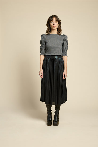 A woman wearing the Feyer Skirt by Cokluch in Glow Knit Black, a midi length pleated skirt in shimmery metallic fabric. She is wearing it with the Insomie top in Facet Black and is standing in front of a beige background. 