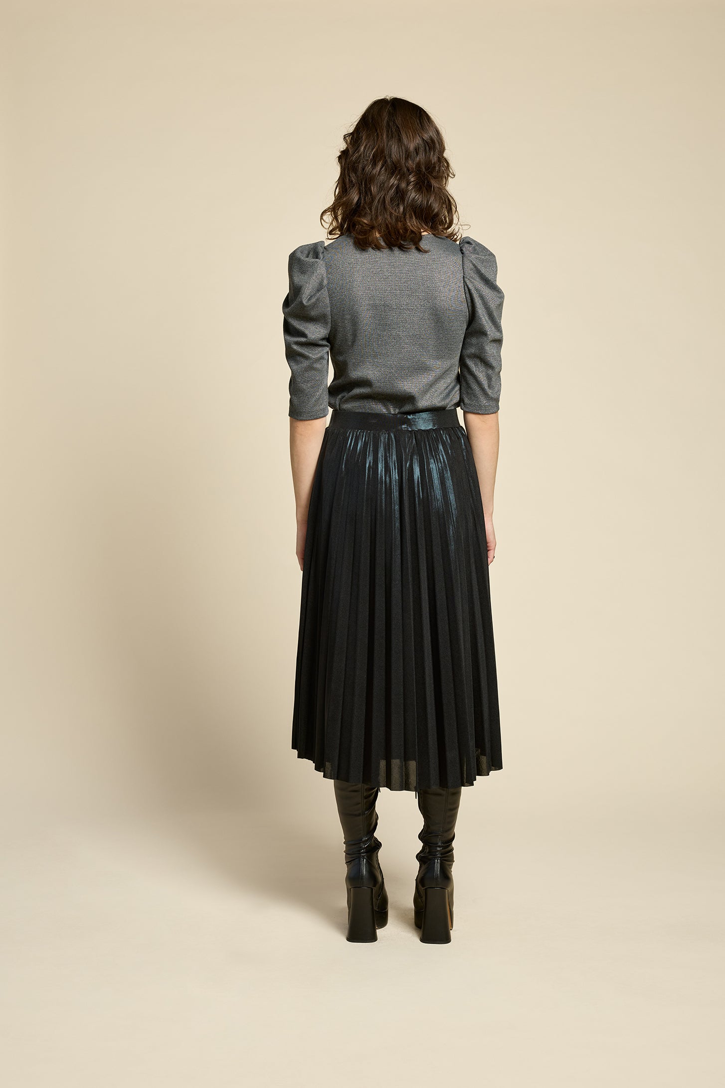 Back view of a woman wearing the Feyer Skirt by Cokluch in Glow Knit Black, a midi length pleated skirt in shimmery metallic fabric. She is wearing it with the Insomie top in Facet Black and is standing in front of a beige background. 